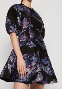 Svart blommig klänning med korta puffärm, prydd med abstrakta mönster i lila, blått och grått. Tillverkad av texturerad tyg med en utsvängd nederkant.