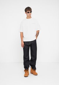 Witte, kort-gewaste t-shirt met grijze opdruk. Donker denim broek en tan werklaarzen met veters en een gestructureerde oppervlakte. Zwarte zonnebril.