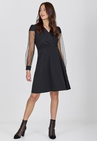 Black dress with a fitted bodice, wrap design, and sheer puff sleeves. Paired with black ankle boots featuring transparent mesh detail.