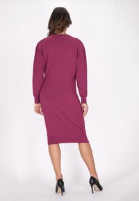 Strickkleid in tiefem Magenta mit einem hinteren Saum bis zum Knie, langen Ärmeln und subtiler Glitzeroptik. Kombiniert mit schwarzen High-Heels.