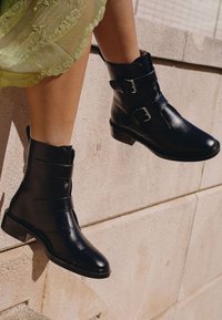 Stivali neri in pelle fino alla caviglia con due fasce dotate di fibbie argentate, punta arrotondata e tacco basso a blocco, su uno sfondo di pietra.