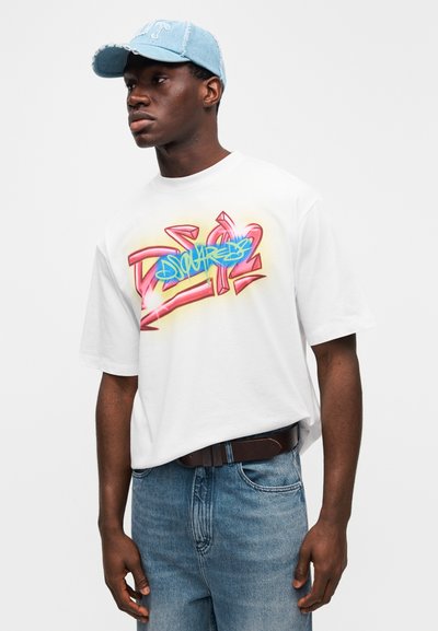 Λευκό υπερμεγέθες t-shirt με πολύχρωμο σχέδιο σε στυλ γκράφιτι· ανοιχτό μπλε τζιν καπέλο· συνδυασμένο με μπλε τζιν παντελόνια και καφέ ζώνη.