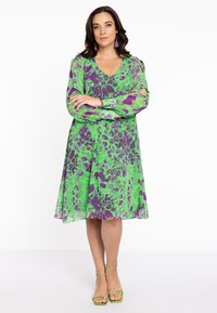 Grünes und lila Leopardenmuster-Kleid mit langen Ärmeln, V-Ausschnitt und A-Linie, kombiniert mit grünen Riemchensandalen. Glatte Stoffstruktur.