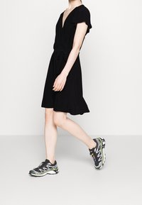 Svart omaxlad klänning med korta volangärmar och knytning i midjan, kombinerad med svarta och gröna sportskor, går på en ljusgrå yta.