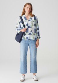 Blusa a maniche lunghe con un motivo geometrico in verde, blu e bianco, abbinata a jeans a campana azzurri chiari e sneakers bianche.