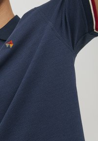 Mornarska polo majica z teksturirano tkanino, kratkimi rokavi in majhnim večbarvnim trikotnim logotipom na levem ovratniku.