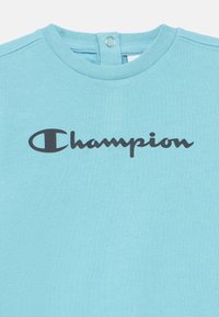 Ανοιχτό μπλε βαμβακερό μπλουζάκι με σκούρο μπλε λογότυπο "Champion" στο στήθος, με κούμπωμα με κουμπιά στο πίσω μέρος του λαιμού για εύκολη εφαρμογή.