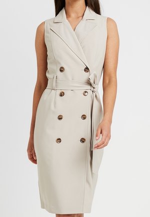 Mouwloze beige double-breasted jurk met reverskraag en taillestrik, gedragen door een vrouw tegen een effen achtergrond.