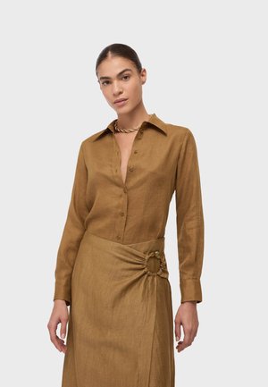Kobieta z zaczesanymi do tyłu włosami, ubrana w brązową, zapinaną na guziki koszulę z długim rękawem oraz dopasowaną spódnicę z marszczonym detalem w postaci pierścienia w talii.