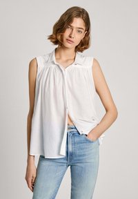Hvid ærmeløs skjorte med knaplukning og plisseret frontdetalje samt små gyldne hardware detaljer, parret med lyseblå denim jeans.
