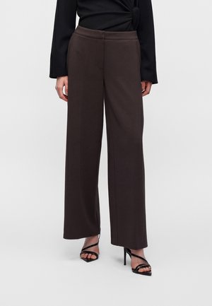 Femme portant un pantalon large marron foncé associé à des sandales à talons noires et un haut noir à manches longues avec un détail noué.