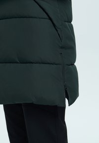 Puffervest i mørkegrøn stof med kviltede sektioner, lynlåslukning i siden og en glat mat overflade mod sorte bukser.