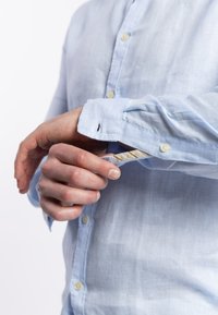 Camicetta azzurro chiaro con bottoni in tessuto traspirante, dotata di polsini a due bottoni e un leggero lucido, con una placca visibile e dettagli interni a contrasto.