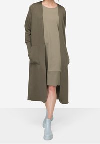 Cárdigan largo verde oliva con dos bolsillos delanteros, superpuesto sobre un vestido beige. Botines de tobillo azul claro con suela gruesa. Textura de tela suave.