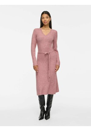 Femme portant une robe en maille rose à manches longues avec un lien à la taille, associée à des bottes en cuir noires montantes jusqu'aux genoux, debout devant un fond blanc.