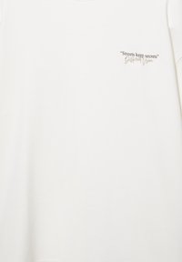 Weißes Hemd mit schwarzem und beigem Text oben links mit den Worten „Streets keep secrets“ und „Different Vision“ in Kursivschrift.