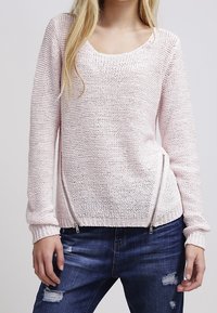 Donna che indossa un maglione lavorato a maglia di un rosa chiaro con due zip diagonali sul davanti, abbinato a jeans scuri strappati.