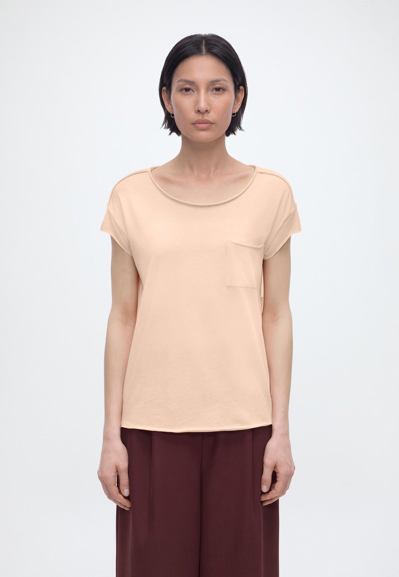 Γυναίκα με κοντά σκούρα μαλλιά, που φοράει μπεζ μπλούζα με κοντά μανίκια και τσέπη στο μπροστινό μέρος, καθώς και καφέ σκούρο παντελόνι με φαρδιά μπατζάκια, κοιτάζοντας μπροστά σε λευκό φόντο.