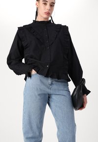 Svart skjorte med knapper som har rynkede detaljer langs fronten og kragen, kombinert med lyseblå denimjeans og en svart håndveske.