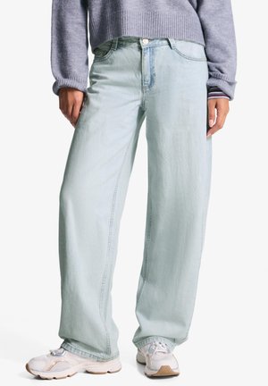 Henkilö, jolla on vaaleansiniset leveälahkeiset farkut, harmaa neule ja valkoiset lenkkarit, seisoo kädet rentoina sivuilla.