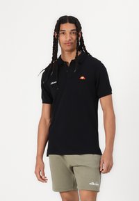 Melna polo krekls ar trīs pogām, ar sarkanu un oranžu logo. Apvienots ar gaiši olīvzaļiem šortiem, ar mīkstu faktūru.