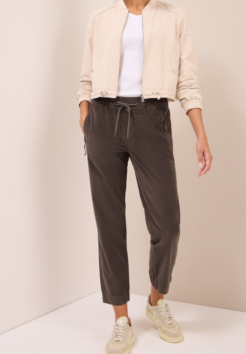 Femme portant une veste beige courte, une chemise blanche, un pantalon gris foncé avec cordon et des baskets beiges, debout contre un mur uni.