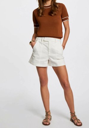 Femme portant un haut en maille marron à manches courtes avec des bordures blanches, un short blanc taille haute, et des sandales plates noires cloutées, les mains dans les poches.