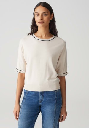 Donna con lunghi capelli scuri che indossa un maglione crema a maniche corte con bordi neri e jeans blu, in piedi contro uno sfondo chiaro e semplice.