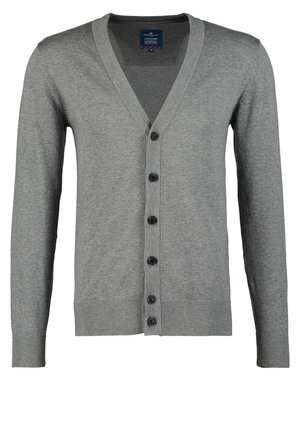 Mann trägt einen grauen, geknöpften Cardigan, weißes T-Shirt, khakifarbene Hose und schwarze Sneaker mit braunen Akzenten, trägt eine braune Ledertasche mit Schulterriemen.
