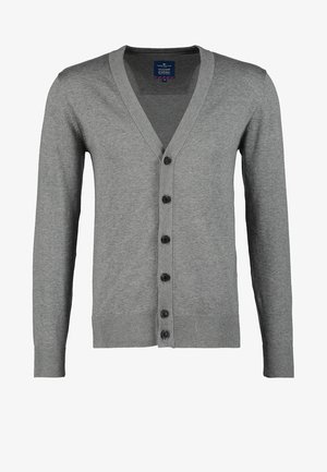 Homme portant un cardigan gris boutonné, un t-shirt blanc, un pantalon kaki et des baskets noires avec des accents marron, portant un sac en cuir marron à bandoulière.
