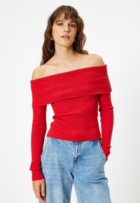 Top rosso a spalle scoperte in maglia a coste con maniche lunghe, caratterizzato da un design aderente e un tessuto testurizzato. Abbinato a jeans azzurri a vita alta.