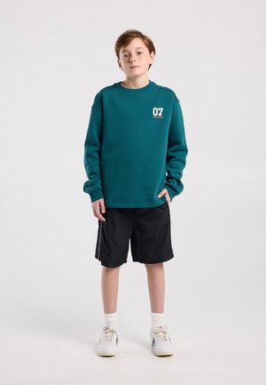 Jongen staat tegen een effen achtergrond, draagt een teal sweatshirt met "07" logo, zwarte korte broek, witte sokken en witte sneakers.