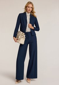 Traje navy a medida con pantalones de pierna ancha y una blazer ajustada. Presenta una blusa rayada de cuello alto y un suave bolso beige.