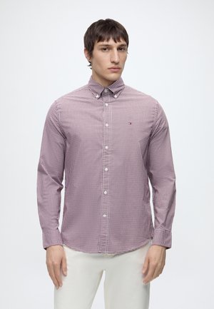 Homme portant une chemise à manches longues à carreaux rouges et blancs boutonnée et un pantalon blanc, debout devant un fond blanc uni.