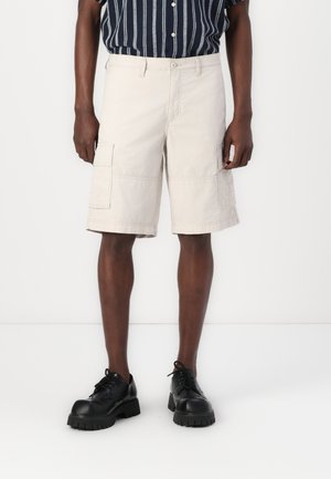 Beige cargo shorts with side pockets, made of lightweight fabric. Worn with a black and white striped short-sleeve shirt and black shoes.