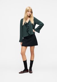 Jeune femme debout, main sur la hanche, portant un chemisier vert à volants, une jupe noire, des chaussettes noires et des ballerines marron sur fond blanc.