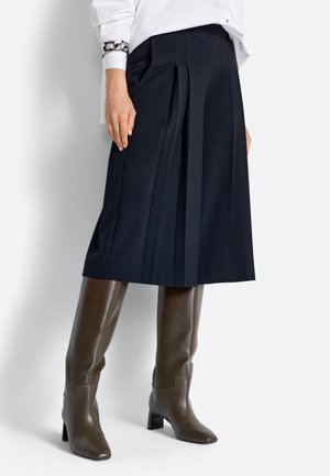 Nő tengerészkék pliszírozott midi szoknyában, barna térdig érő magas sarkú csizmával, fehér blúzzal és díszes karkötővel.