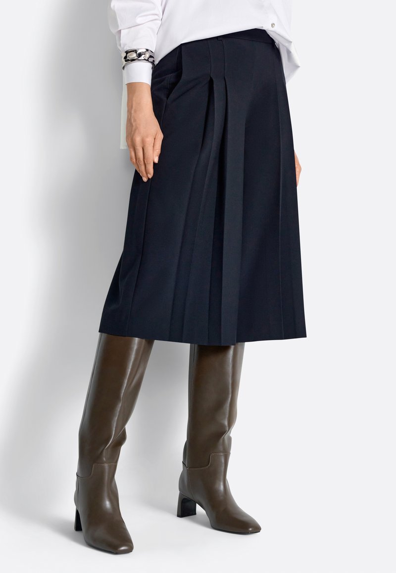 Moteris vilki tamsiai mėlyną klostuotą midi sijoną, rudus aukštakulnius aulinukus iki kelių, baltą palaidinę ir dekoratyvinį apyrankę.