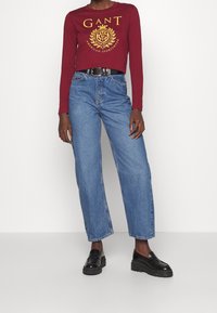 Rotes Langarmhemd mit goldenem Logo, kombiniert mit lockeren blauen Jeans. Schwarze Plateauschuhe runden das Outfit ab.