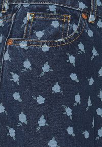 Tejido de mezclilla en azul oscuro con un patrón floral que presenta rosas azul claro y hojas verdes. Costuras en contraste y un detalle en el bolsillo trasero.