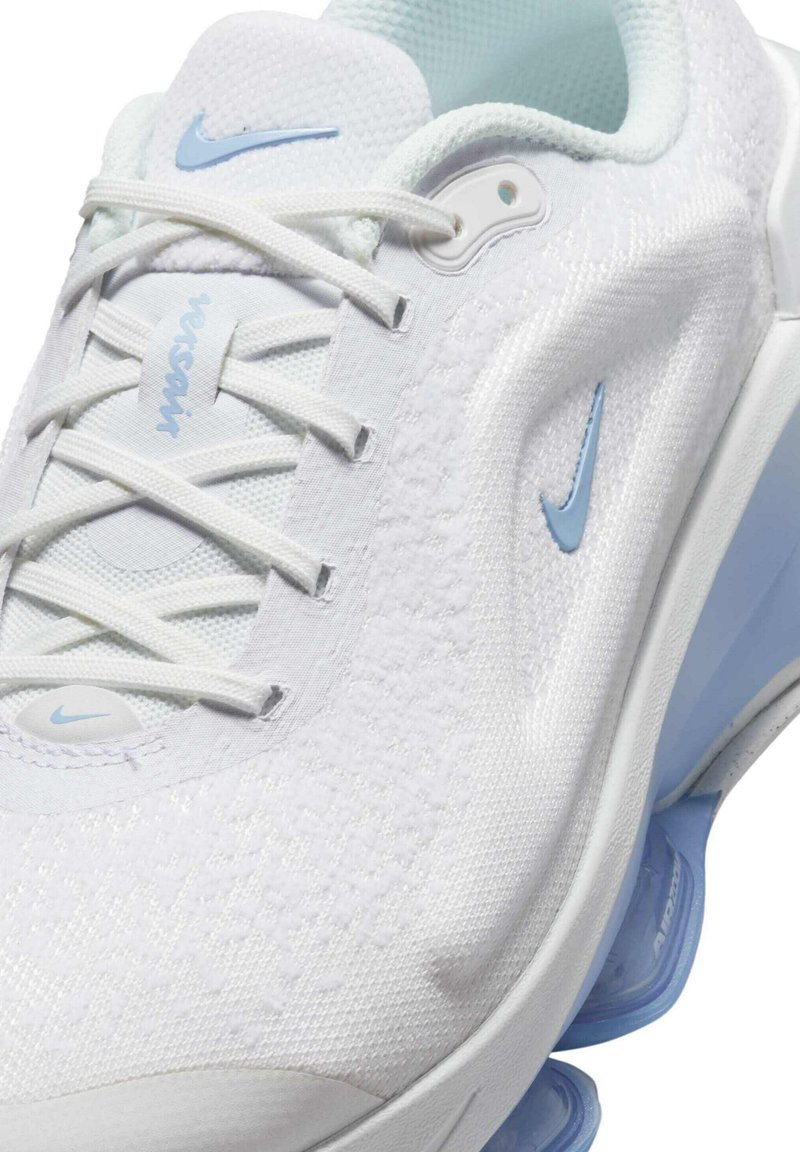 Scarpa sportiva bianca con tomaia texturizzata, dettagli azzurri chiari, chiusura con lacci e unità di ammortizzazione visibile nel tallone. Presenta il marchio Nike.