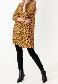 Gul klänning med svart blommönster, trekvartslånga ärmar, lös passform, ihop med svarta smala byxor och anklar stövlar. Enkelt design.