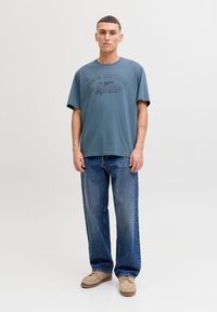 Dark blue cotton t-shirt with embossed text, relaxed fit, accompanied by light blue jeans and beige moccasin shoes. Neutral background.