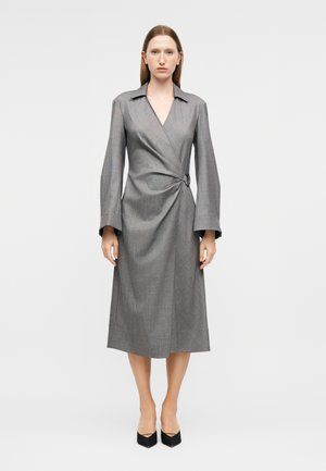 Frau trägt ein graues Wickelkleid mit langen Ärmeln, einem Kragen und einer seitlichen Schnallendetail, kombiniert mit schwarzen spitz zulaufenden High Heels.