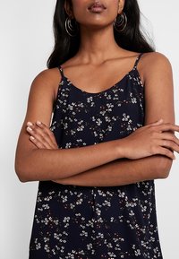 Žena se středně hnědou pletí, oblečená v námořnicky modrých květovaných šatech bez rukávů a zlatých kruhových náušnicích, se založenýma rukama na hrudi.