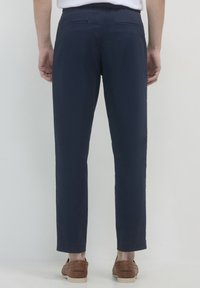 Homme portant un pantalon slim bleu marine et des mocassins en cuir marron se tient contre un fond clair, dos à la caméra.
