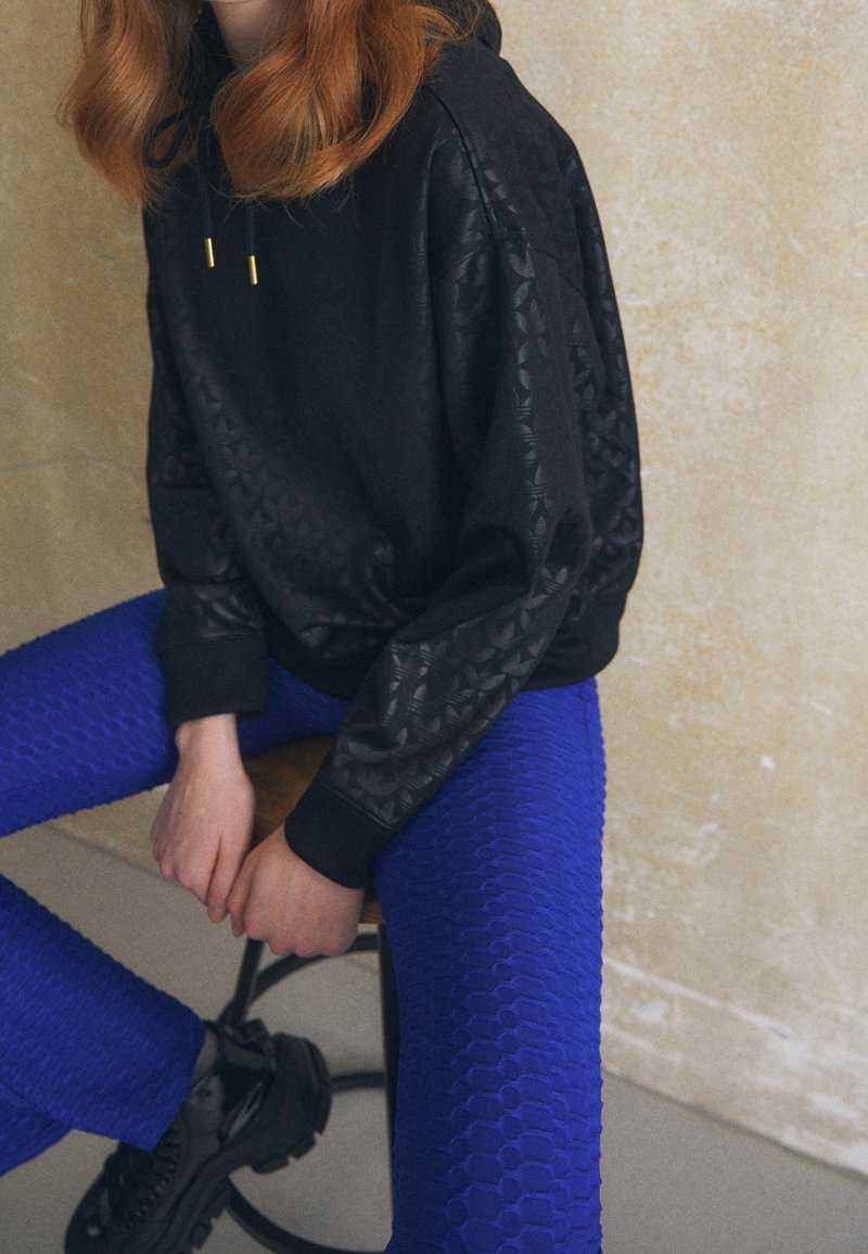 Sudadera negra con mangas estampadas, combinada con mallas brillantes azul claro de textura que presentan un diseño hexagonal y zapatos negros de suela gruesa.