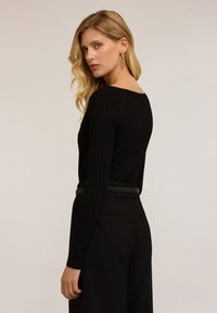 Donna bionda con capelli lunghi e ondulati, che indossa orecchini a cerchio dorati, un top nero a maniche lunghe a costine e pantaloni neri a vita alta su uno sfondo neutro.