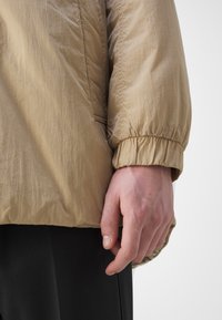 Lichte, beige jas met een gestructureerde afwerking; aansluitende manchetten; zichtbare stiksels en naaddetails; gedragen over zwarte stof.