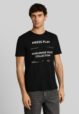 Junger Mann mit lockigem Haar, trägt ein schwarzes T-Shirt mit weißem Text "PRESS PLAY WORLDWIDE MUSIC COLLECTION" und dunkelgraue Jeans, stehend.